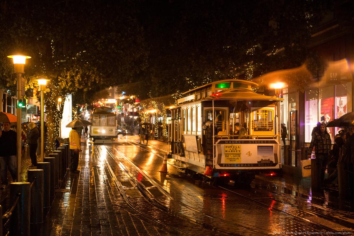 Cable Car 4 travels down wet Powell Street at night, decorated with holiday lights and garland.