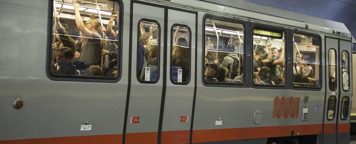 Crowded Muni Metro train car with passengers standing and holding overhead rails.