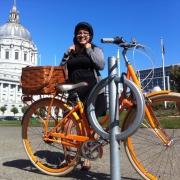 Woman in black jacket adjusting helmet on orange bike with Capitol building in background.