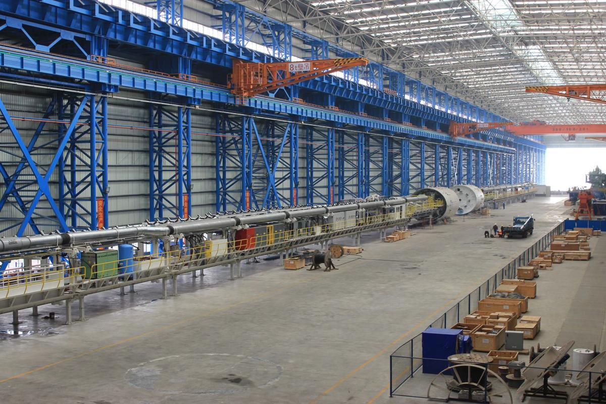 Large tunnel boring machine in industrial warehouse with blue overhead cranes and support structures.