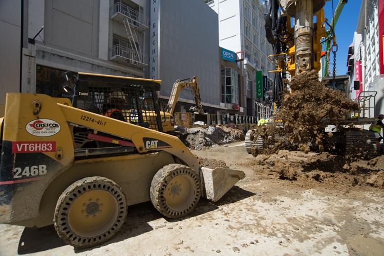 Construction equipment removing excavated soil on an urban street with buildings in background.