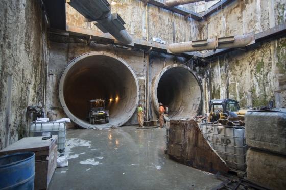 Two large circular concrete tunnels with construction equipment and workers visible inside an underground construction site.