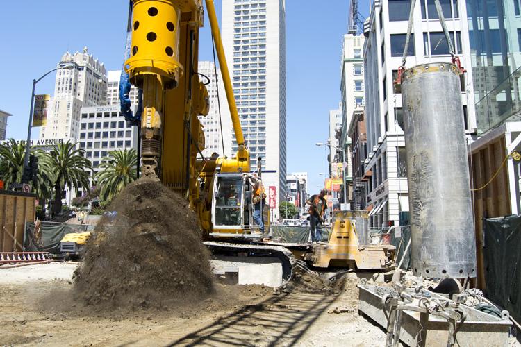 Yellow excavator digs at construction site on urban street with high-rise buildings and palm trees.