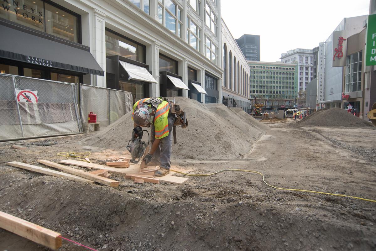 Construction worker in safety vest building wooden forms on urban street surrounded by buildings and equipment.