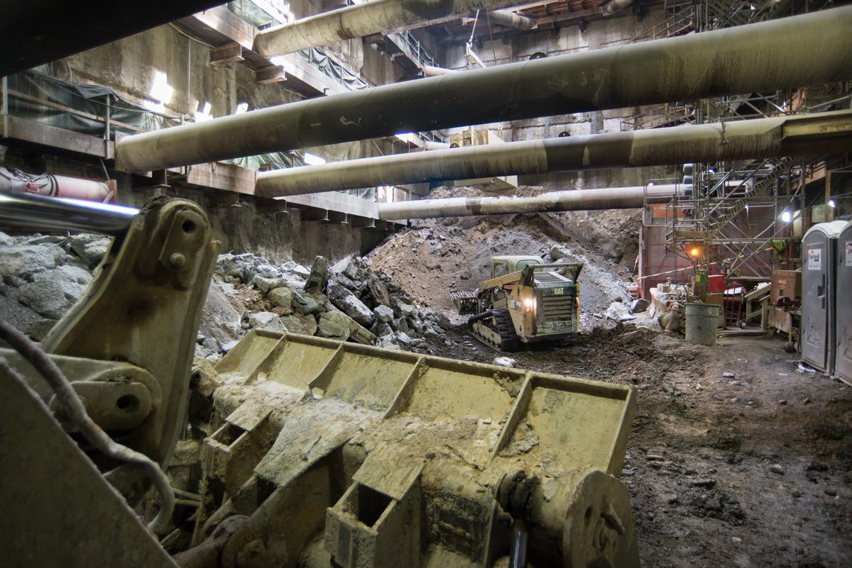 Construction equipment excavating underground platform cavern with large pipes overhead and concrete structures above.