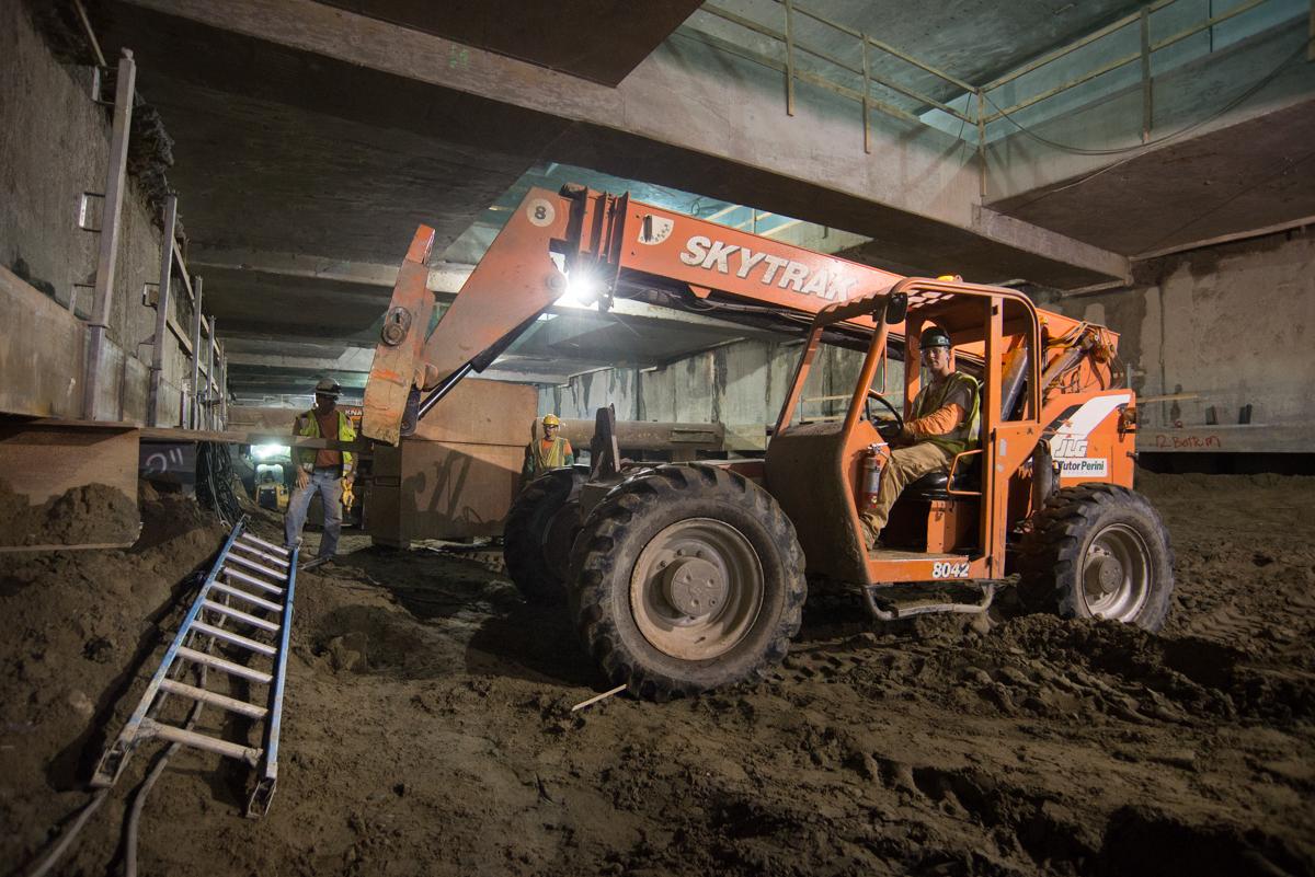 Crew members operate an orange Skytrak forklift to move materials inside an underground station construction area.