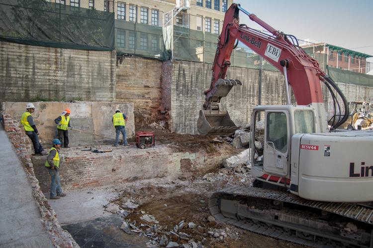 Excavator with red boom removing debris from demolished building site, workers in yellow vests supervising urban construction area.