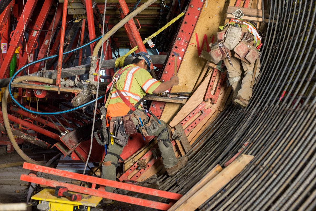 Worker in safety vest and harness adjusting formwork and reinforcing steel for concrete pour in underground transit station construction.
