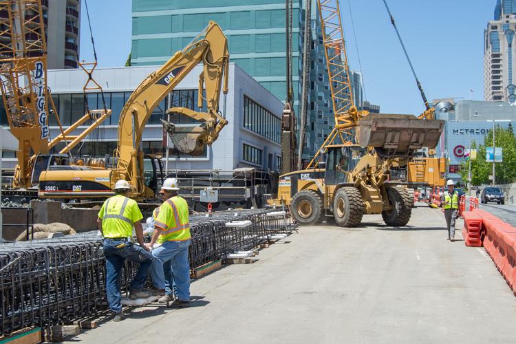 Construction site with excavator, wheel loader, and workers in safety vests on urban street surrounded by tall buildings.