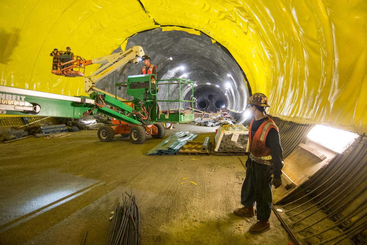 Workers in safety gear install waterproofing on tunnel exterior using boom lifts and equipment inside yellow-lined tunnel.