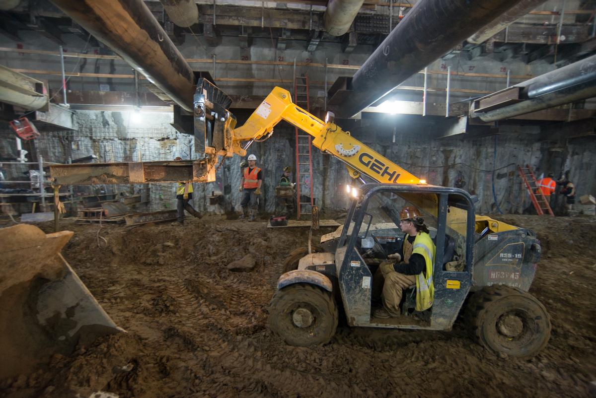 Construction equipment operates inside underground transit station during restoration work, with workers and steel beams visible.