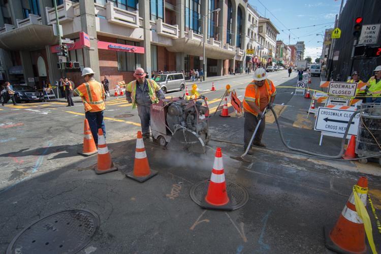 Construction workers in safety vests operate street maintenance equipment on a closed urban street with traffic cones and sidewalk closure signs.