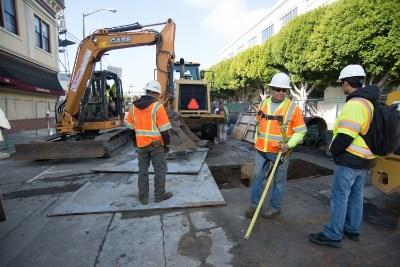 Workers in safety gear perform potholing work to locate underground utilities at a street intersection.