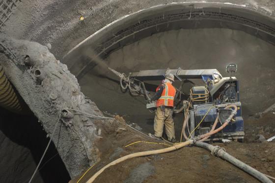 Worker in safety vest spraying shotcrete inside underground tunnel construction for Central Subway project.