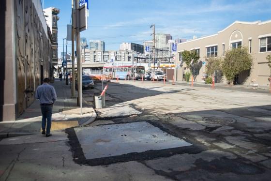 Urban street with construction site, damaged pavement, and city skyline in background.