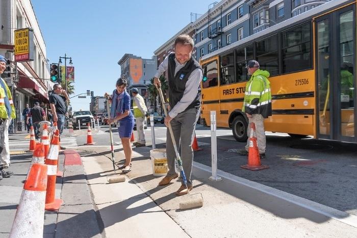 Workers paint a pedestrian safety zone on a city street near a yellow school bus and traffic cones.