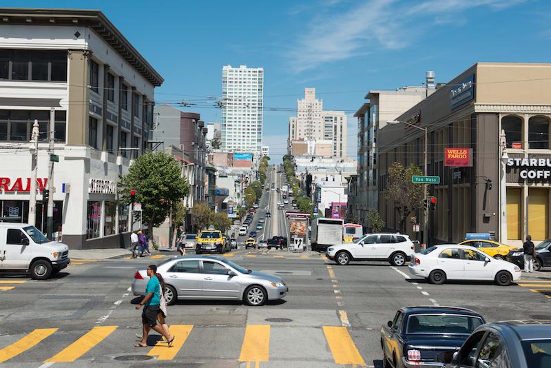 Busy intersection with cars, taxis, bus, and pedestrians crossing at Van Ness Avenue in San Francisco.
