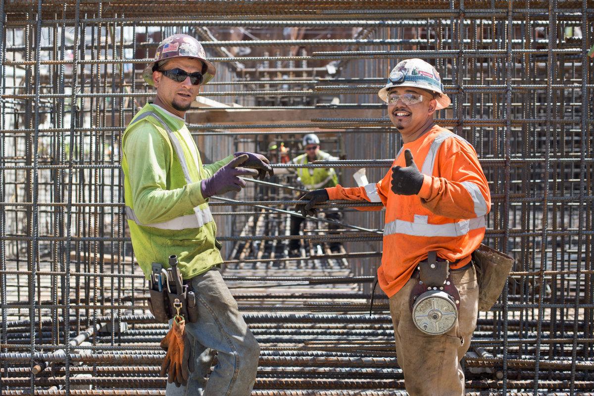 Two construction workers in safety vests and hard hats stand before steel rebar framework at a transit station construction site.