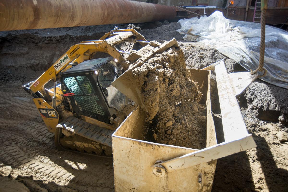 Yellow skid steer loader filling steel box with excavated soil at transit station construction site.