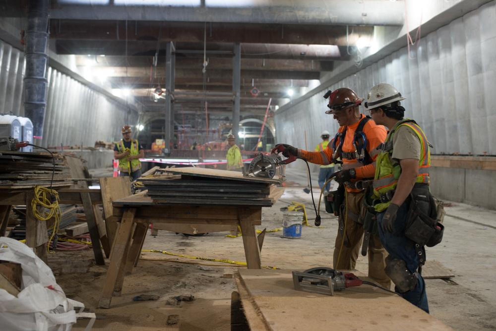 Workers in hard hats and safety vests score plywood sheets for concrete formwork at a Central Subway construction site.