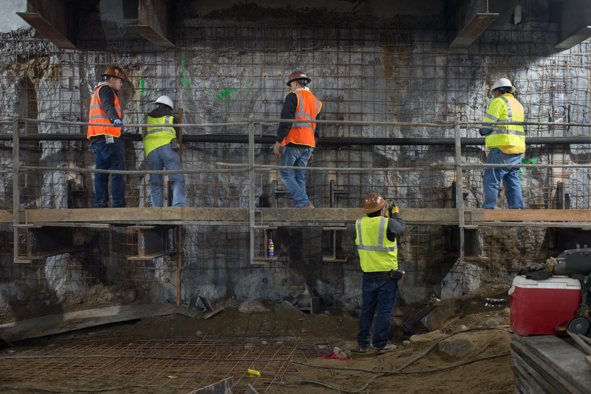 Construction workers in safety vests and hard hats inspect steel reinforcement mesh and install conduit on an underground station excavation wall.
