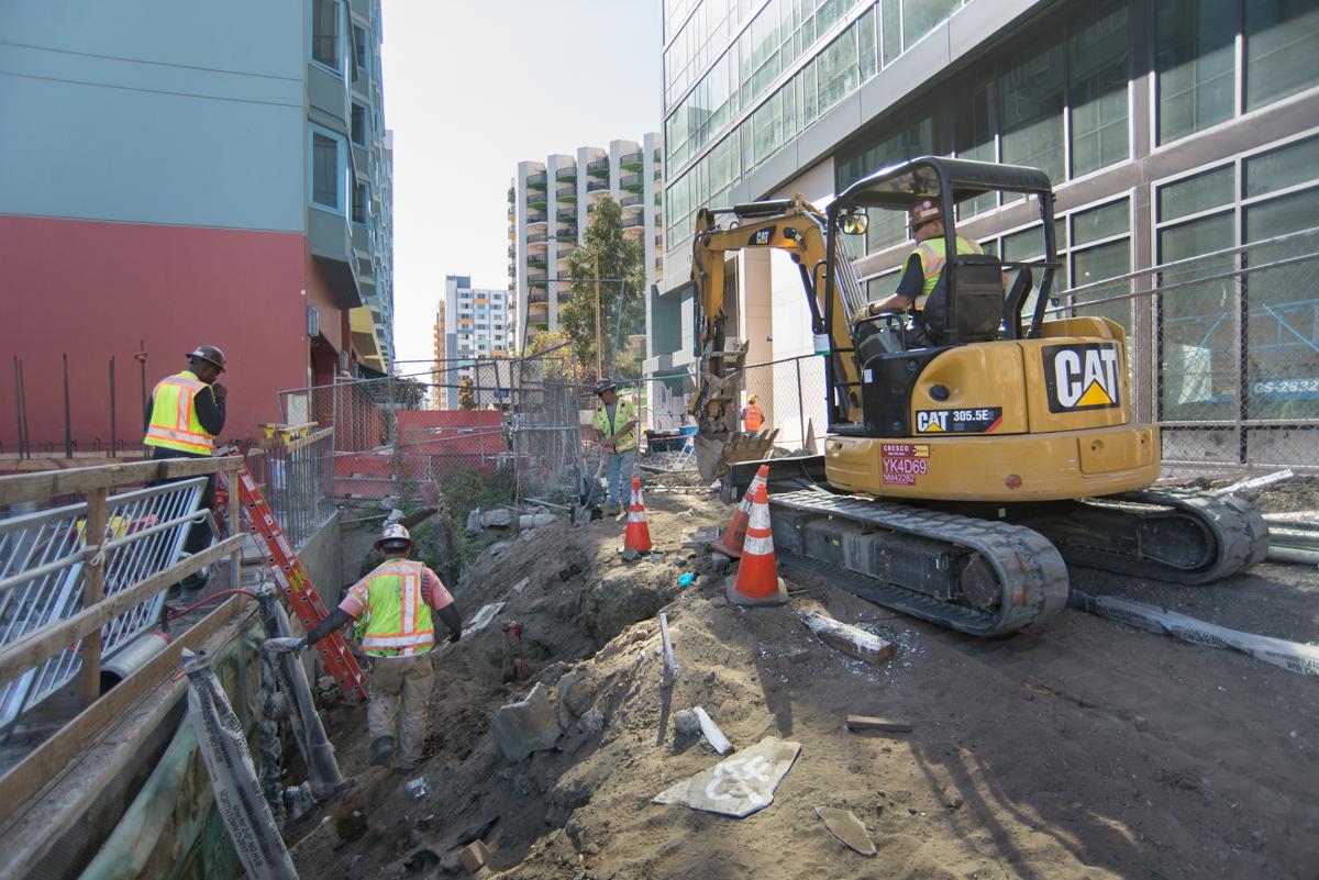 Construction crew operates excavator while installing utilities on urban street with surrounding buildings.
