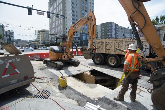 Construction crew operates excavator at street work site near 4th and King Streets for Central Subway utility upgrades.