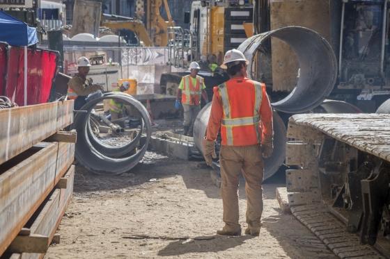 Construction workers in safety vests handle large concrete pipes at an urban transit construction site.