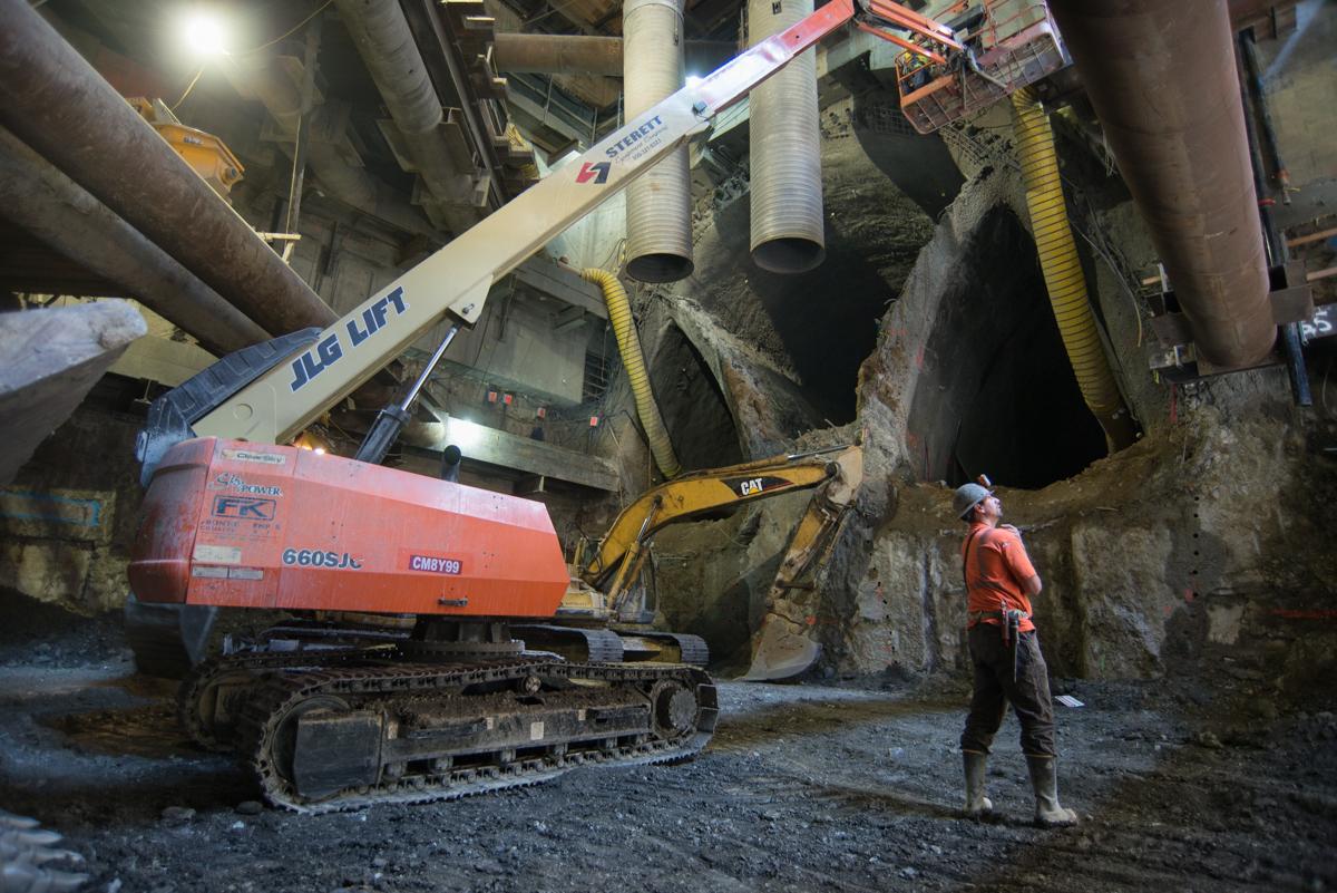 Excavation equipment and worker at underground transit station construction site with concrete pipes and structural elements.