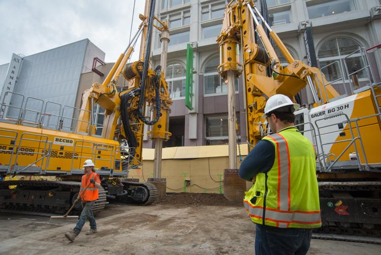 Construction crew operates yellow auger drill equipment on urban street near building facade.