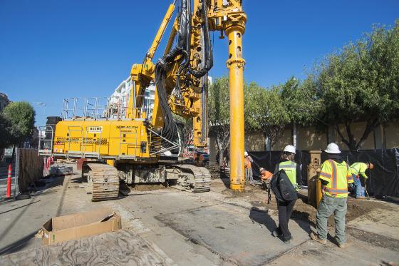 Yellow drilling rig performs foundation work on city street with construction workers and trees visible.