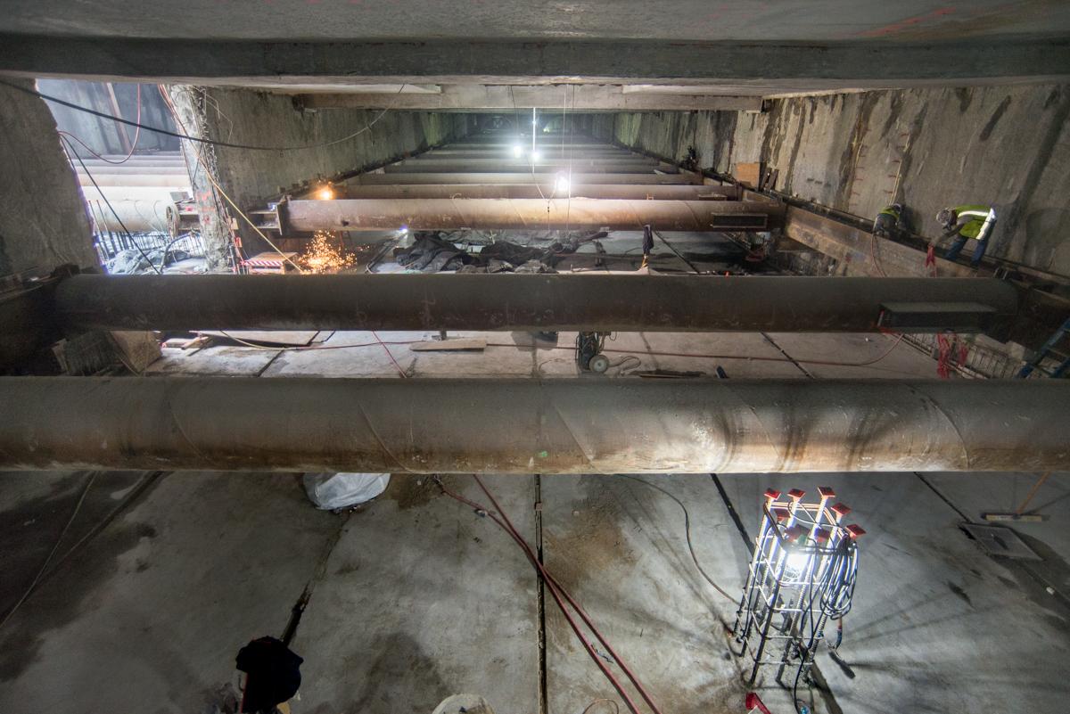 Construction underway inside underground transit station box with concrete structural elements and work lights.