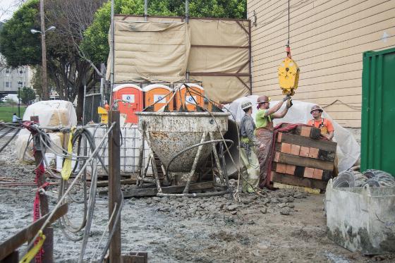 Construction workers operate concrete mixer and equipment at street work site with yellow hoist and building materials.