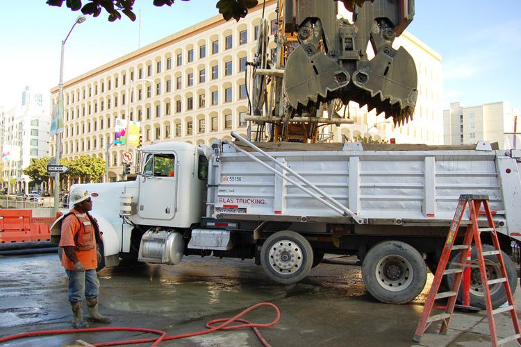 Worker stands beside dump truck with excavator overhead on wet street in urban area.
