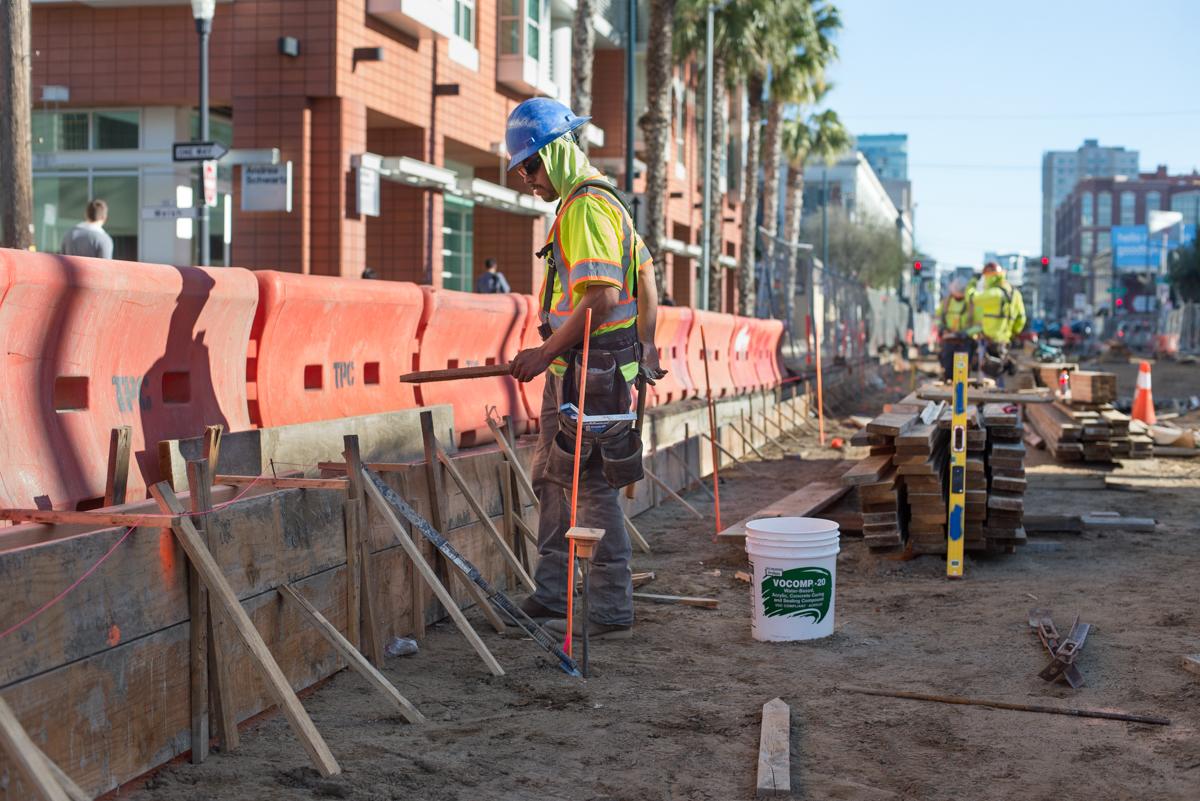 Construction worker in safety gear and hard hat operates jackhammer on 4th Street, with orange barriers and wooden forms visible along active transit project site.