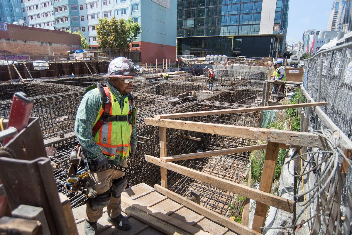 Construction worker in safety gear at Yerba Buena/Moscone Station site with rebar cages and wooden framing visible.