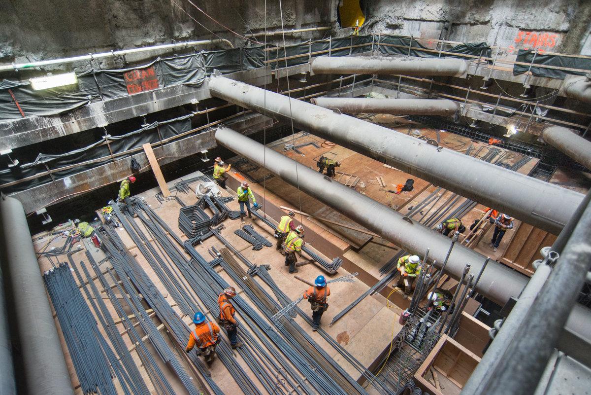 Construction workers assemble rebar at bottom of underground transit station excavation site.