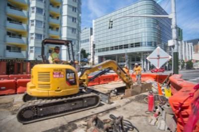 Yellow excavator and construction crew working on 4th Street with traffic cones and barriers visible.