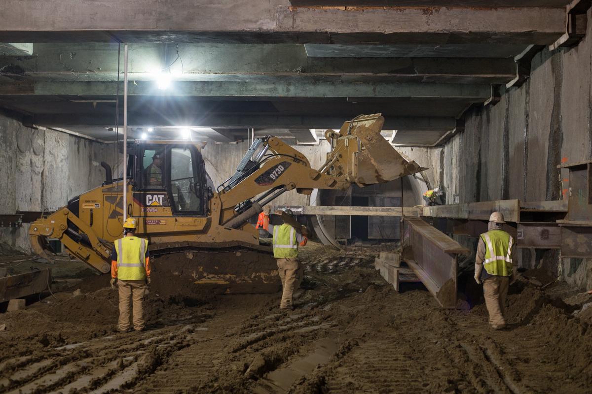CAT excavator loads soil into truck inside underground station box with workers in safety vests observing.