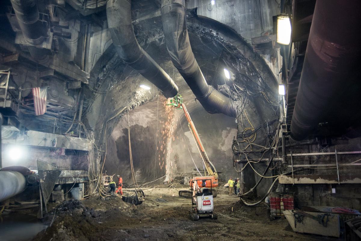 Workers use equipment to modify ventilation infrastructure inside underground transit tunnel cavern with overhead pipes and lighting.