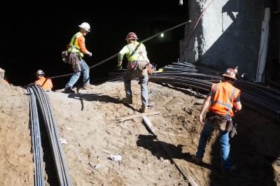 Construction workers in safety gear excavating a trench for transit station box frame structure.