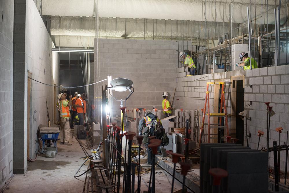 Construction workers in safety gear build interior walls and install steel framing inside a transit station under bright work lights.