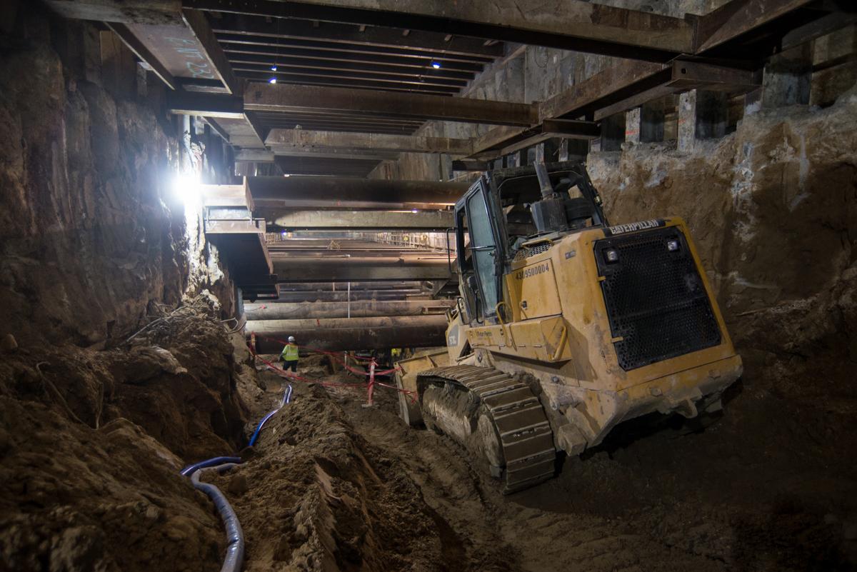 Yellow bulldozer removing excavated material from underground station construction site beneath city streets.
