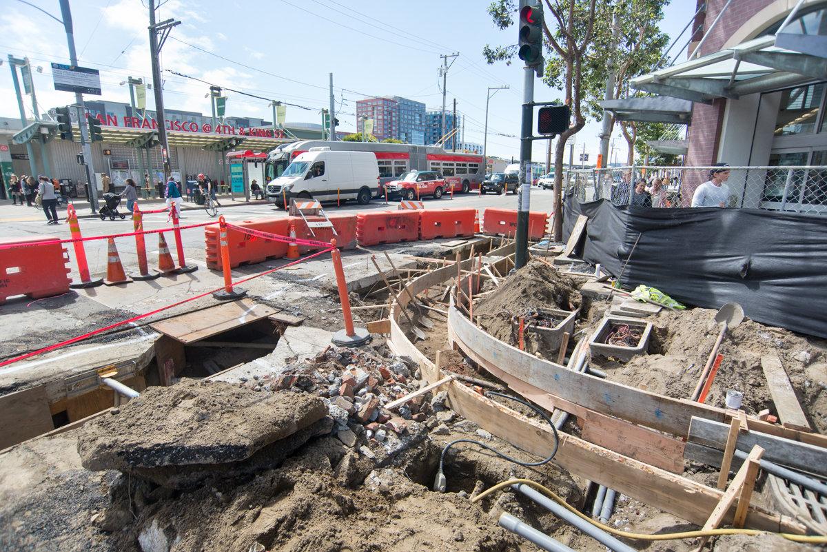Construction site on 4th Street with excavation, concrete forms, and red safety barriers along urban street.