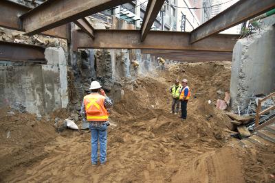 Workers in safety vests excavating soil beneath concrete beams at a transit construction site.