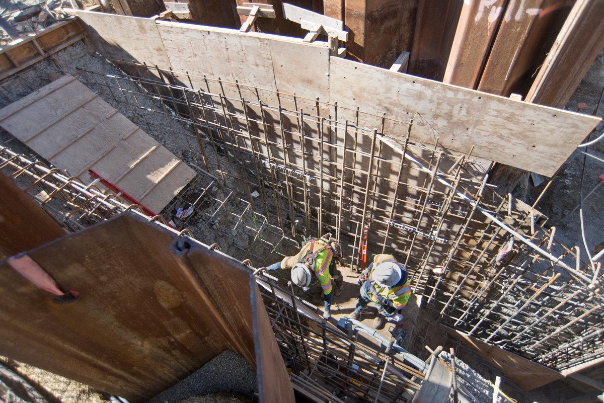 Two workers in safety gear assemble steel reinforcement and scaffolding inside a station construction pit.