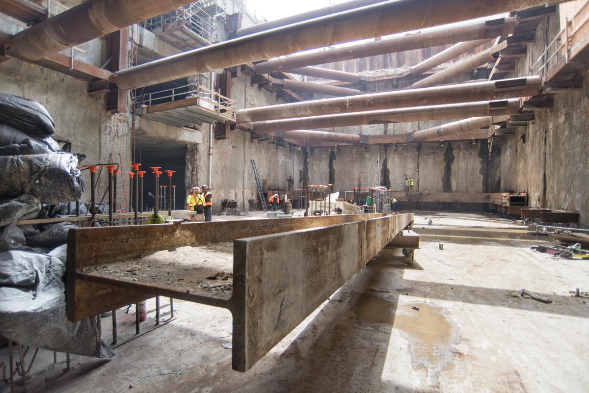Industrial construction site with concrete structural elements, steel beams, and workers in safety gear inside a large underground station box.