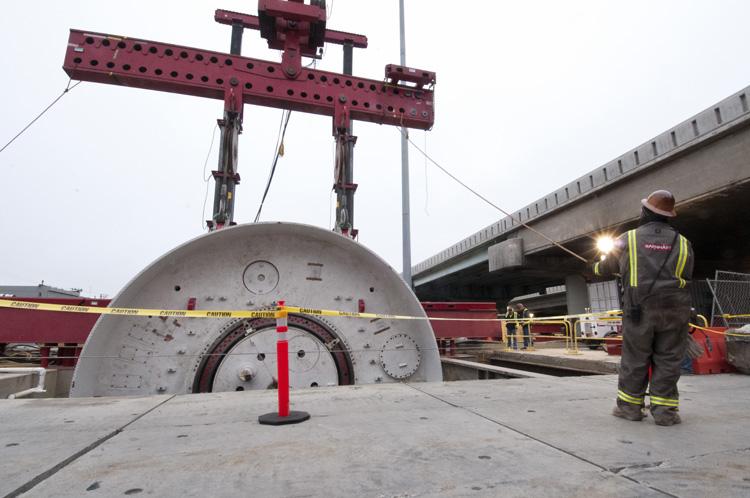 Large tunnel boring machine segment being lowered by red industrial crane at underground construction site with worker nearby.