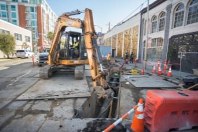Excavator digging trench on urban street with orange safety cones and construction barriers.