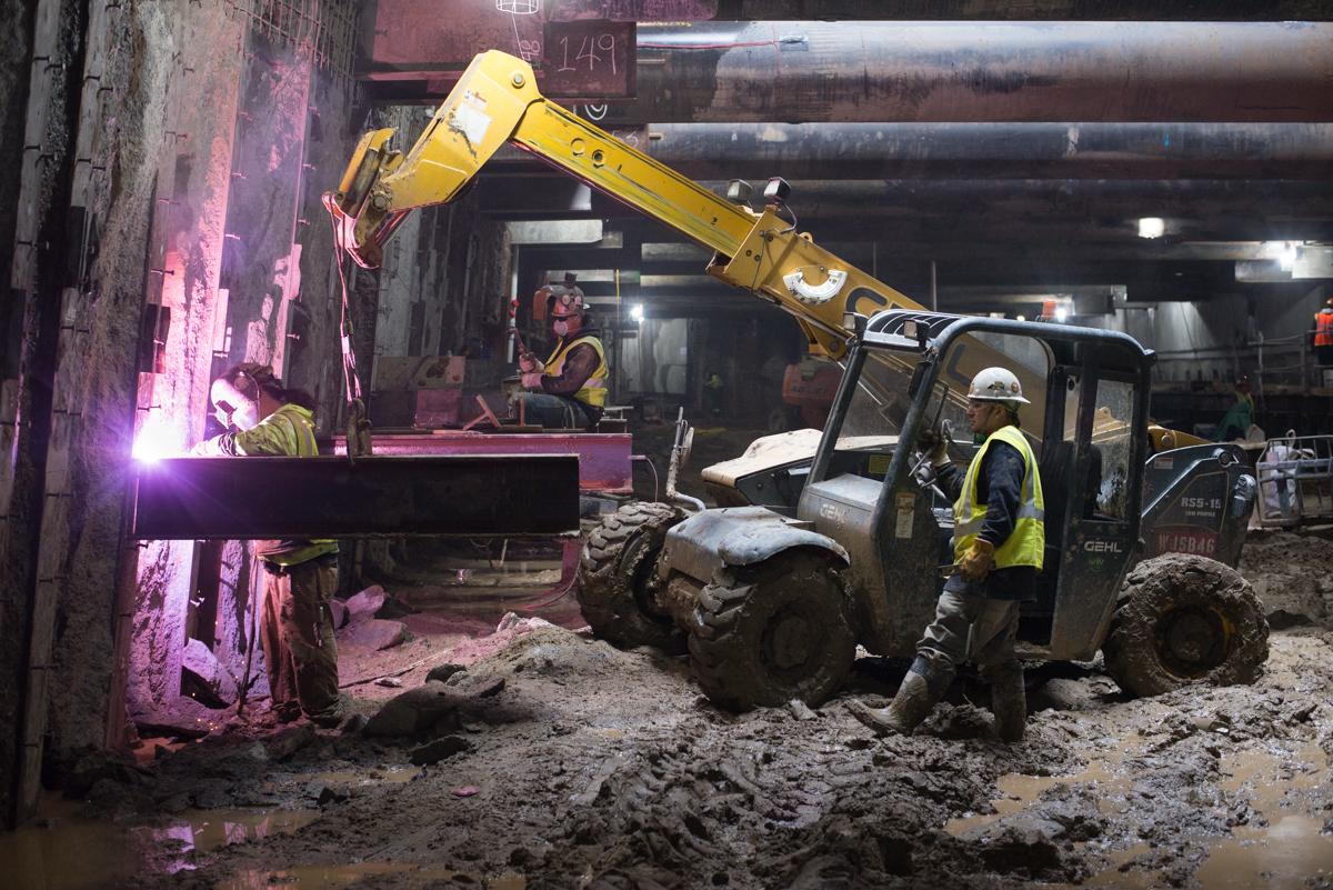 Workers weld a steel beam at night using heavy equipment on a construction site.